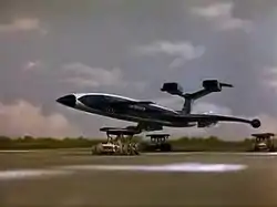 A blue, futuristic airliner rests on top of three orange support vehicles (one underneath the fuselage and two underneath the wings) as it speeds out of control down an airport runway, the sky bright with clouds.