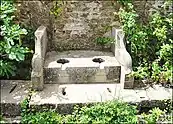 A stone bench-like construction with two holes in it, in front of a brick wall. There are plants growing around it.
